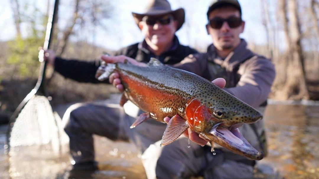 Two men fishing in a river. The man on the left holding a net and the man on the right holding a trout