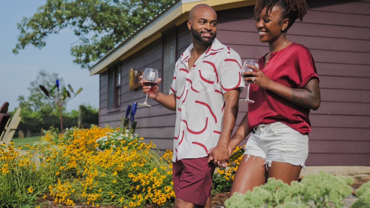 A couple holds each other's hands and glasses of wine while strolling through a vineyard in Blue Ridge, GA